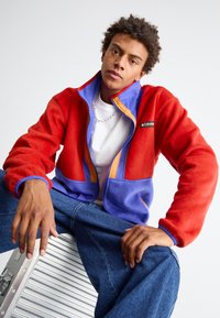 Giacca in fleece rossa e viola con chiusura a zip, colletto alto e dettagli arancioni a contrasto. Modello che indossa jeans blu, seduto.