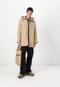 Beige vandtæt jakke med hætte, frontlommer, en grå inderside, parret med sorte cargo-bukser og en beige tote-taske. Brune sko.