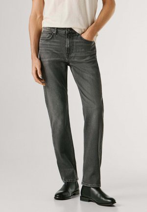 Uomo con jeans neri dritti sbiaditi, camicia bianca e stivali Chelsea in pelle nera, in piedi davanti a uno sfondo chiaro e uniforme.