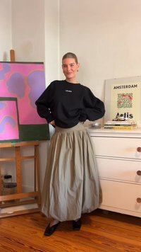 Črn oversized pulover s napisom "LEGER", v kombinaciji z dolgo, nabrano khaki krilom. Model nosi črne čevlje na barvitem ozadju.