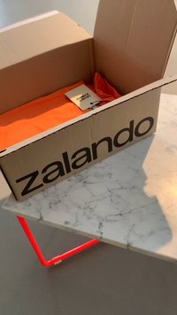 Kartonny pārvadājumu kaste ar "zalando" logo, kurā ir oranža auduma gabals un birka. Novietota uz marmora galda ar sarkanu metāla rāmi.