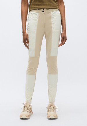 Beige bukser med et tætsiddende design, der har kontrasterende paneler og lynlåslommer. Fremstillet af et let, glat stof.