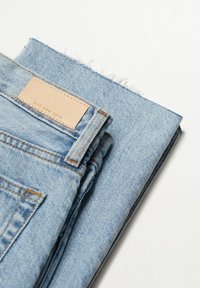 Jean en denim bleu clair délavé avec une finition texturée, plié soigneusement. Présente une étiquette en cuir sur la ceinture avec un texte embossé.