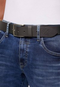 Schwarzer Ledergürtel mit strukturiertem Finish, ausgestattet mit einer Metallschnalle; getragen über blauen Denim-Jeans mit kontrastierenden Nähten.