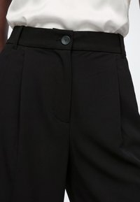 Pantalon noir taille haute avec fermeture par boutons, porté avec une chemise blanche rentrée, mettant en valeur la taille et la partie supérieure de la cuisse.