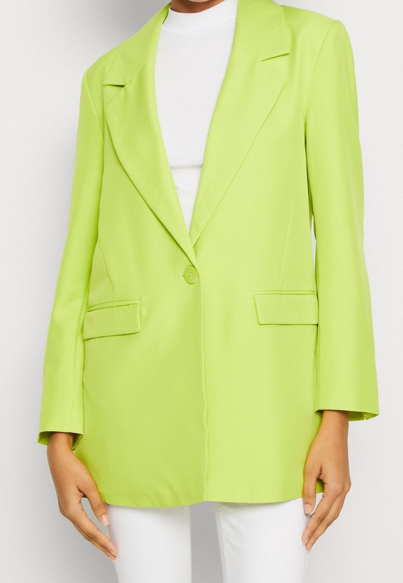 Lime green blazer valmistettu sileästä kankaasta, jossa on yksittäinen nappikiinnitys, viistekaulukset ja kaksi etutaskua. Muodikkaasti oversized-malli.