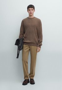 Brązowy dzianinowy sweter, beżowe spodnie, czarna skórzana torba w ręku, ciemne buty, neutralne tło, luźny krój i prosty design.