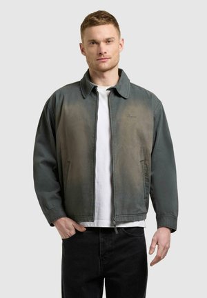 Mann som står med den ene hånden i lomma, iført en falmet grønn jakke med glidelås over en hvit t-skjorte og svarte bukser mot en nøytral bakgrunn.