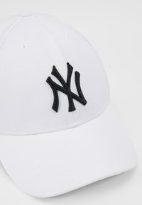 Hvid baseballcap lavet af tekstureret stof med et fremtrædende sort broderet logo på forsiden. Struktureret form med en buet skygge.