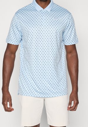 Man draagt een lichtblauw poloshirt met korte mouwen met kleine donkere patronen en witte shorts, staand tegen een effen achtergrond.