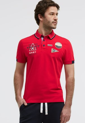 Mann trägt rotes Polohemd mit marineblauem Saum und maritimen Aufnähern, steht mit Händen in den Taschen und schaut zur Seite.