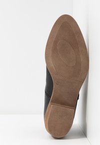 Chaussure en cuir noir avec un bout lisse et arrondi, dotée d'une semelle en caoutchouc beige. L'intérieur affiche une taille 37. Design simple avec des accents minimalistes.