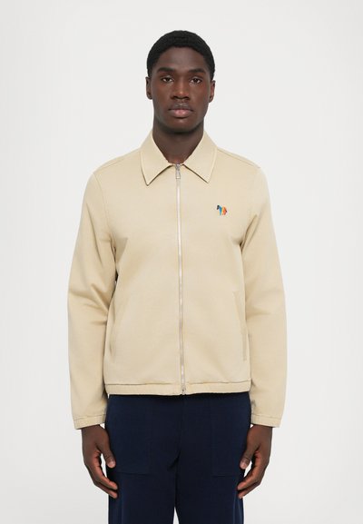 Giovane uomo che indossa una giacca beige con zip e un logo colorato di cavallo sul petto, con pantaloni blu scuro, in piedi davanti a uno sfondo bianco uniforme.