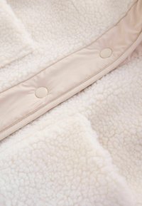 Gros plan sur un tissu sherpa doux de couleur crème avec une bordure beige clair et deux boutons-pression ronds le long de la couture.