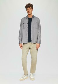 Giovane uomo in piedi, che indossa una camicia grigia chiara sopra una maglietta nera, pantaloni beige e sneakers bianche su uno sfondo chiaro e semplice.