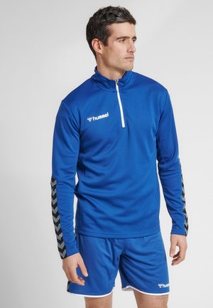 Blaues sportliches Viertelreißverschlusshemd mit strukturiertem Finish, schwarze Chevron-Musterakzente an den Ärmeln, kombiniert mit passenden blauen Shorts.