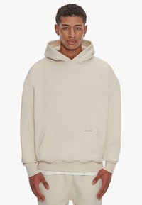 Beige Kapuzenpullover mit strukturierter Oberfläche, im Oversized-Fit und mit Fronttasche. Besitzt einen gewellten Saum und ein aufgenähtes Etikett.