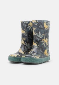 Une paire de bottes de pluie sombres avec des semelles vertes, ornées d'un motif de lionceau de dessin animé jaune et de feuillages jungles verts sur un fond blanc.