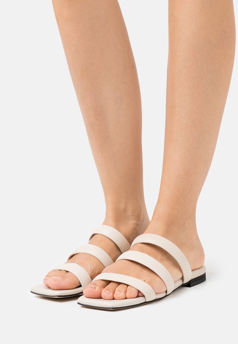 Sandali piatti beige chiaro con quattro fasce larghe, design open-toe e una suola nera bassa. Texture liscia con uno stile minimalista.
