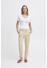 Beige mönstrade byxor med sidfickor, tillsammans med en vit figurnära topp och bruna platta sandaler. Mjukt tyg med en subtil textur.