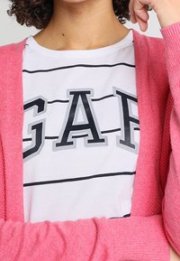 Růžový kardigan přes bílou košili s černými vodorovnými pruhy a výrazným šedým logem "GAP" v tučných písmenech. Měkké textury.