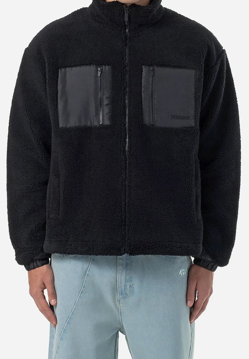 Mężczyzna ubrany w czarną polarową kurtkę z dwiema dużymi przednimi kieszeniami na zamek i jasnoniebieskie dżinsy.