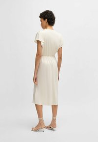 Abito in crema fluente con maniche corte a volant, vita elasticizzata, lunghezza al ginocchio e texture liscia abbinato a sandali con cinturini di colore chiaro.