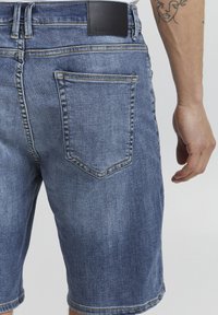 Pantalones cortos de denim en azul claro con una textura suave. Presentan una etiqueta de cuero negro en la cinturilla, costuras dobles y un bolsillo trasero.