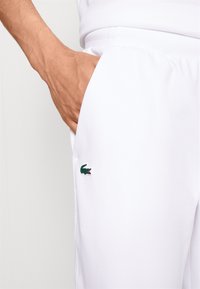 Hand i fickan på vita byxor med en liten broderad grön krokodil-logotyp på vänstra låret.