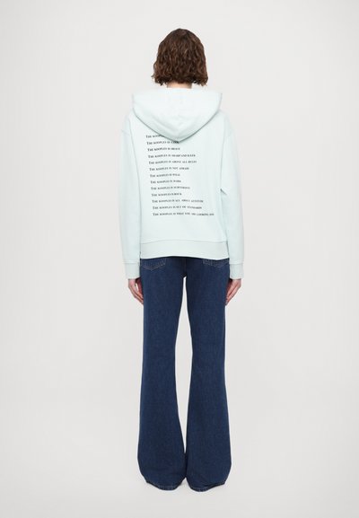 Hoodie v meti zeleni barvi z natisnjeno besedilo na hrbtni strani, v kombinaciji z razširjenimi temno modrimi kavbojkami. Mehka tkanina, sproščen kroj in nastavljiva kapuca.