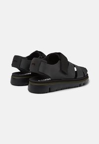Black sandal shoes with adjustable straps, rubber soles, and a textured finish, featuring cut-out details for breathability and comfort.