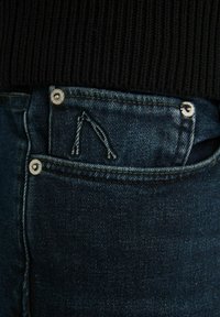 Mörka denimjeans med en texturerad tyg, prydda med silverfästen och en distinkt broderad logotypdetalj nära fickan.