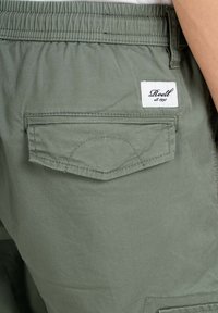 Pantalones cortos verde caqui con cintura elástica, un bolsillo trasero y una etiqueta blanca con texto negro cosida en la parte trasera.
