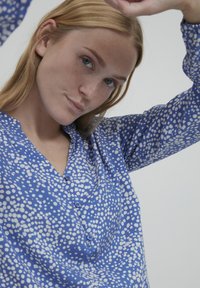 Blusa blu con motivo a pois bianchi, chiusura frontale con bottoni e una texture in tessuto morbido. Maniche lunghe con una leggera lucentezza.