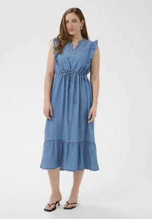 Kobieta w niebieskiej, midi sukience bez rękawów z falbanami na ramionach i dolnej krawędzi, stojąca w czarnych sandałach na obcasie na jednolitym białym tle.