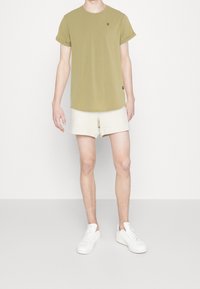 Olivově zelené tričko s krátkým rukávem a kulatým výstřihem, se malým logem, v kombinaci se světle béžovými šortkami a bílými teniskami.