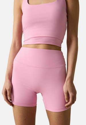 Femeie îmbrăcată într-un top roz fără mâneci și pantaloni scurți asortați, cu talie înaltă și croială mulată, pe un fundal simplu.