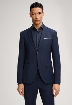 Uomo che indossa una giacca di abito blu scuro e pantaloni abbinati sopra una camicia blu abbottonata, in piedi contro uno sfondo chiaro e uniforme.