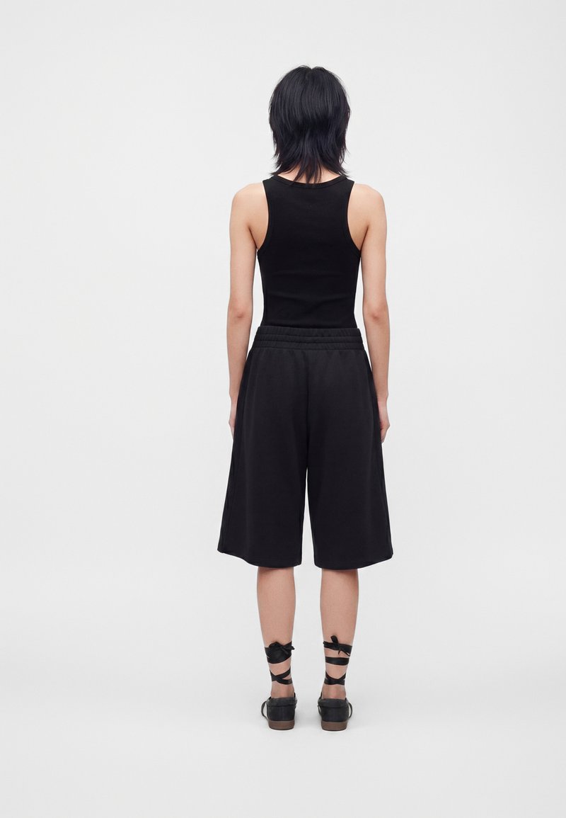 Personne aux cheveux courts noirs portant un haut noir sans manches, un short noir ample jusqu'aux genoux et des chaussures noires avec des brides à la cheville, debout de dos.