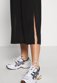 Schwarzes Midi-Kleid mit Seitenschlitzen, aus weichem Stoff; getragen mit weißen und marineblauen Sportschuhen mit orangefarbenen Akzenten und Schnürsenkeln.