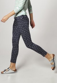 Kalhoty s námořnickým vzorem a bílými geometrickými motivy, doplněné metalickými stříbrnými sandály a zeleno-bílým pruhovaným tričkem.