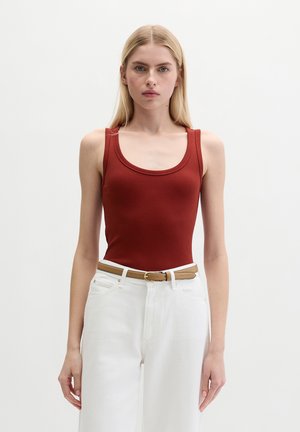 Vrouw met steil blond haar, gekleed in een roestkleurige tanktop, witte hooggetailleerde broek en een beige riem, staand tegen een effen achtergrond.