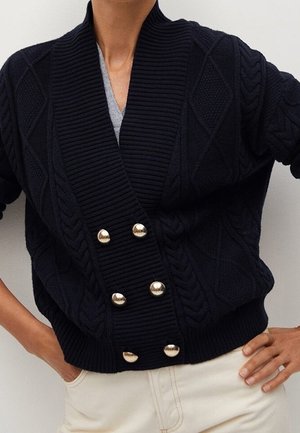 Osoba w czarnym kardiganie z grubym splotem i dużymi złotymi guzikami na szarym topie, do tego jasne spodnie, ręce na biodrach.