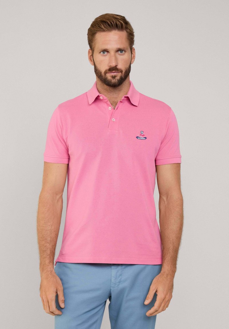 Maglietta polo rosa con colletto e due bottoni, caratterizzata da un piccolo logo ricamato blu e bianco sul lato sinistro del petto. Maniche corte.