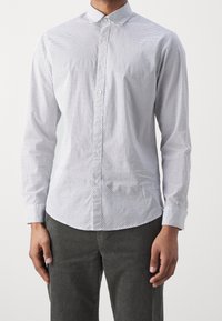 Camicia bianca con bottoni, decorata con pois neri, maniche lunghe e colletto classico. Il tessuto appare liscio con una leggera lucentezza.