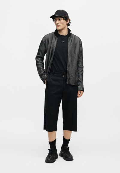 Νέος άνδρας που φοράει μαύρο ριγέ μπουφάν με φερμουάρ, μαύρο καπέλο, φαρδιά μαύρα κοντά παντελόνια, μαύρες κάλτσες και χοντρές μαύρες αθλητικές παπούτσια μπροστά σε απλό φόντο.