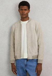 Helebeež suede bomber jaak ribakudumisetega mansetide ja äärtega, lukuga ning küljetaskutega, paarituna valge ümarakaelusega sviitriga ja siniste teksadega.