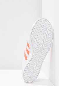 Vit sneaker med orange ränder, med en texturerad sula och ett mjukt ovandel-material. Synlig varumärkeslogotyp på sidan.