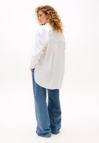 Camicia bianca con bottoni e piega sul retro, abbinata a jeans in denim azzurri a gamba larga. Tessuto morbido e vestibilità rilassata.