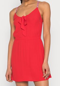 Červené šaty bez rukávů s mašlí vpředu, tenkými ramínky s logovými akcenty a volnou, mírně rozšířenou sukní.